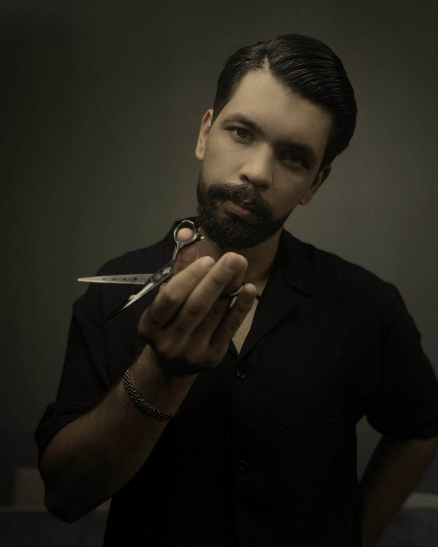 A barber with slicked-back hair and a beard holds a pair of scissors against a dark backdrop.