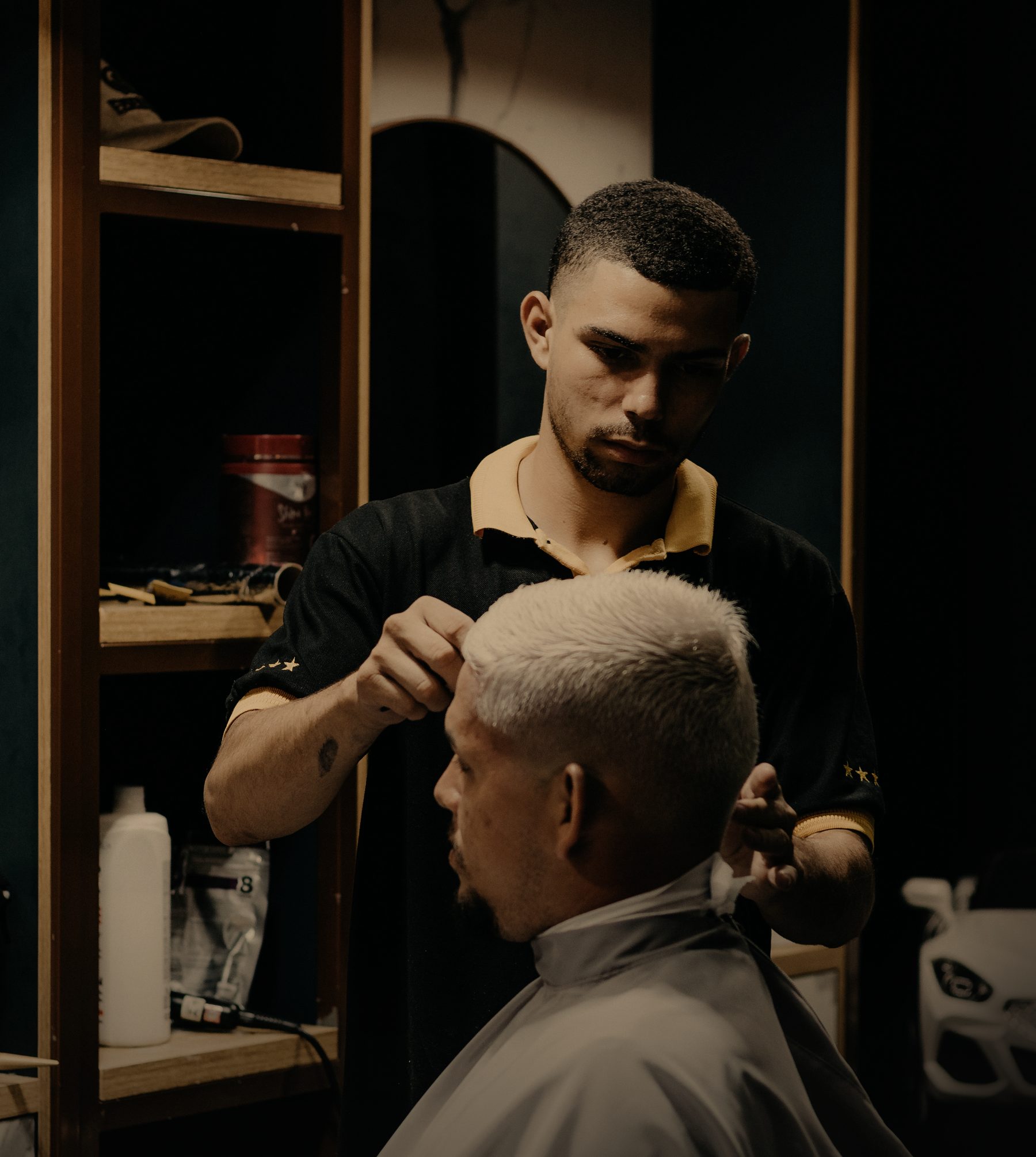 A barber in a dark polo shirt shapes a client's bleached crop in a warmly lit shop.