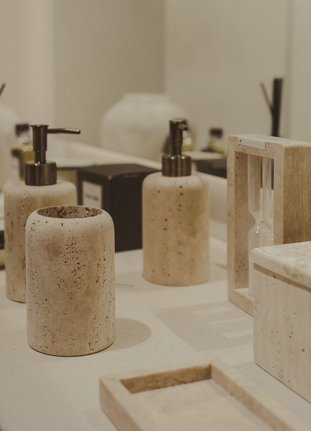 Travertine soap dispensers, a tumbler, an hourglass, and a tray on a pale stone counter.