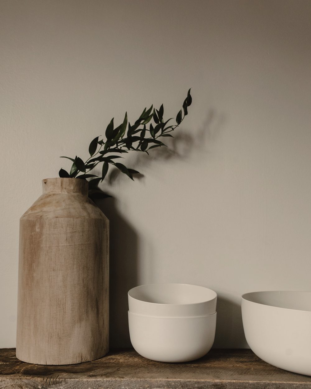 A wooden-toned vase with a green sprig and two white bowls on a shelf against a pale wall.