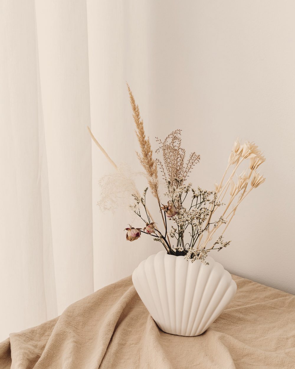Dried flowers in a white shell-shaped vase on draped beige linen.